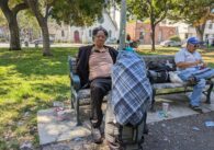 An unhoused man sits on a bench in a park