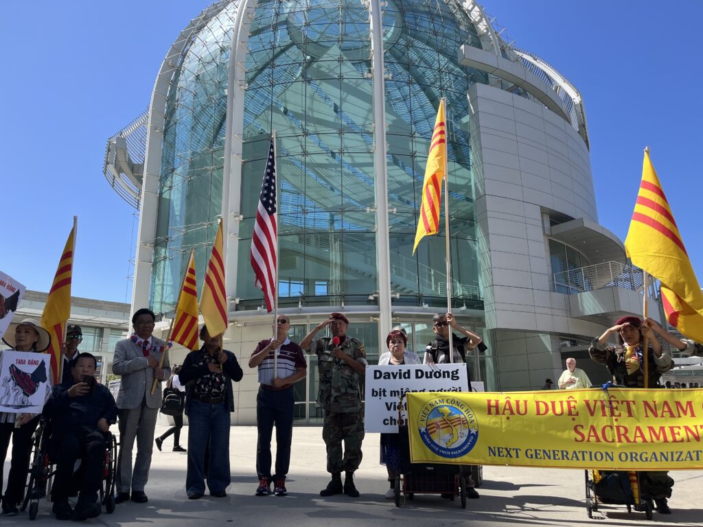A group of people holding flags and signs protest in front of San Jose City Hall