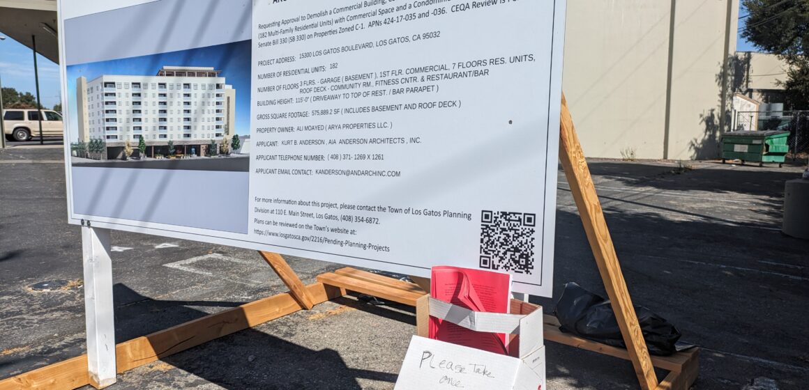 A notice of development proposal sign with a homemade cardboard box holder carrying red flyers attached to the sign.