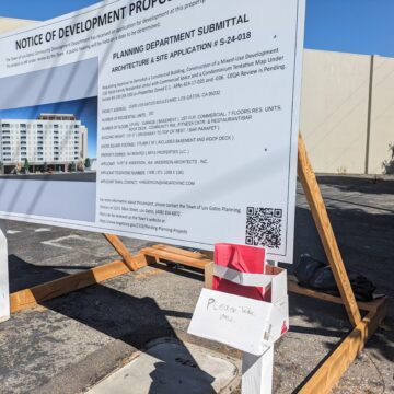Un cartel de aviso de propuesta de desarrollo con un soporte de caja de cartón hecho en casa que lleva folletos rojos adheridos al cartel.