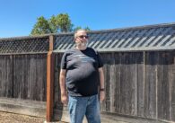 A middle-aged man wearing glasses and a black t-shirt stands in front of his backyard fence.