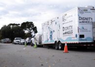 Multiple cars parked along the street, including the Dignity on Wheels mobile shower and laundry unit, which has three cones in front of it and the truck that carries it
