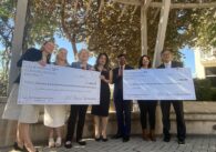 A line of people stand outside holding two oversized checks