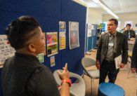Man in suit with grey tie and visitor pass stands in education space talking with person in foreground