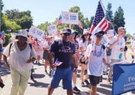 Group of people walking holding American flags and signs reading "Assemblymember Evan Low", centered on a man wearing an Oakley shirt, a hat, and sunglasses