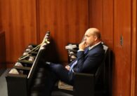 Man in blue suit sits and watches something out of frame with an empty row of seats in front of him