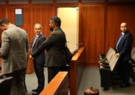 Three people standing in front of a barrier talking with someone out of frame while one man in a suit stands behind barrier and behind a row of seats