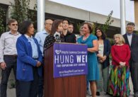A woman stands behind a lectern with microphones surrounded by people.