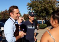 Patrick Ahrens, a light skinned man with brushed back light brown hair, wearing a brown apron over a blue button down, standing talking with supporter with their brown hair in a bun wearing a spaghetti strap top