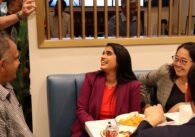 Tara Sreekrishnan, brown skinned woman with long, straight black hair, wearing a purple blazer and red shirt, seated and looking up while talking with a supporter out of frame