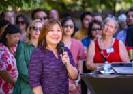 A middle-aged woman stands holding a microphone speaking in front of a crowd.