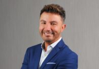 A man in a blue suit jacket stands with his arms crossed smiling at the camera.