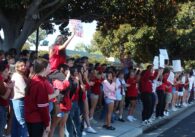 A large group of high school students wearing red stand along a curb at school holding signs that read things like, "Our teachers deserve more than praise. They deserve a raise!"