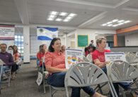 A group of people sit in rows of grey chairs with signs that read "Keep LGBTQ students and teachers safe at CUSD"