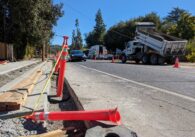 Orange bollards line a trench dug for new sidewalk along a road with a Tesla driving. Construction trucks sit parked across the street.