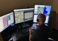 A man sits at a desk, holding a controller and facing four compute screens with maps on them.