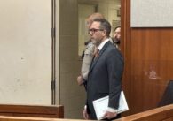 A man in a suit stands in court, with the bailiff and an incarcerated man behind him