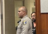 A correctional officer stands in front of an incarcerated man in court