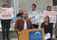 A man stands at a podium with people in the background holding signs
