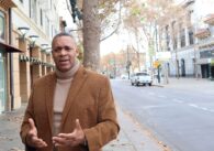 A man in a turtleneck sweater and suit jacket stands outside on the sidewalk next to the street