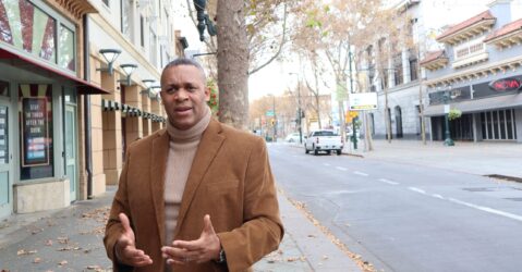 A man in a turtleneck sweater and suit jacket stands outside on the sidewalk next to the street