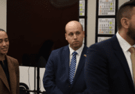 Three people in suits standing, one woman and two men. The man in the middle wearing a striped tie and American flag pin watches something in the distance