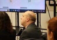Anthony Becker, light skinned, partly bald man wearing a grey suit, sitting with his lawyers in a courtroom