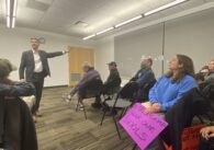 A man stands speaking to a crowd of people in chairs