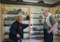 An older man and woman stand holding a historic figurine in front of shelves of figures.