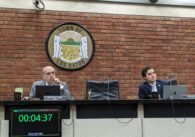 Two men in suits sit behind a council dais with laptops and microphones.