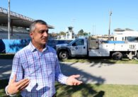 Man wearing plaid shirt talking in front of large pool with construction trucks parked outside