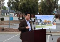 Man wearing brown blazer and button shirt speaking at podium with a board showing the development renderings