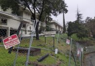 A two-story abandoned building behind a wire fence with a "No trespassing" sign