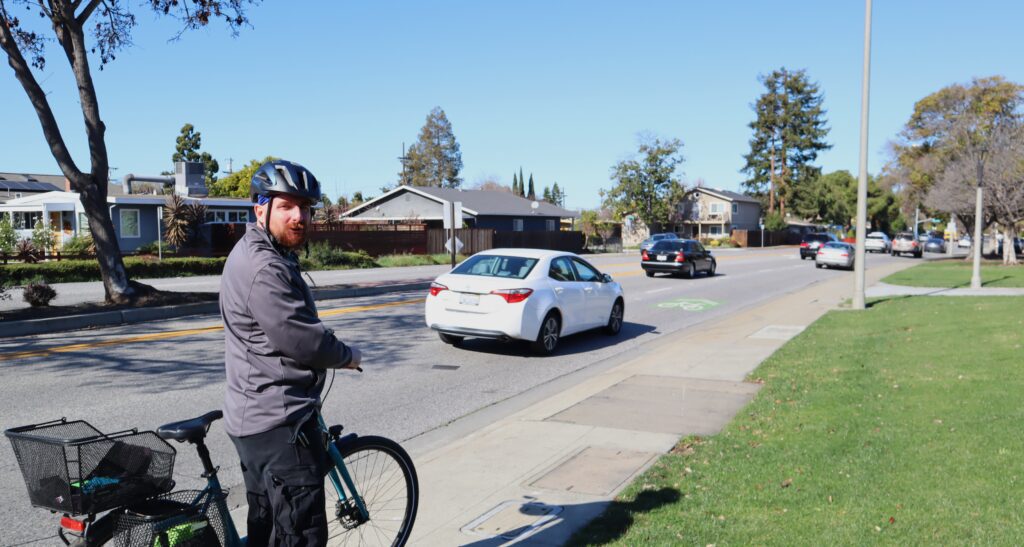 A man stands outside with his bike, with a car parked in the background