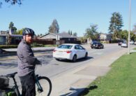 A man stands outside with his bike, with a car parked in the background