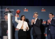Woman wearing white jacket holding football standing on stage beside applauding men