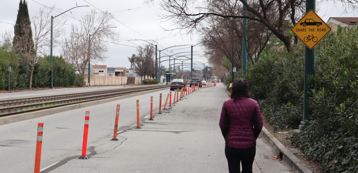 Woman in puffer jacket walks in blocked pedestrian lane while cars drive on the open lanes, with a "share the road" sign above her