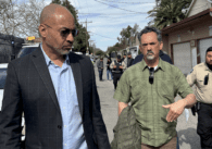 Một người đàn ông đeo kính đang đi bộ cùng một người đàn ông khác trên một con phố dài và đông đúc.