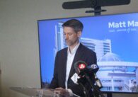 Man standing in front of a blue screen talking in front of a podium with press microphones nearby.