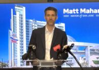 A man in a suit standing in front of a podium of news media microphones in front of a blue screen reading "Matt Mahan, Mayor of San Jose."
