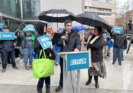 A man stands surrounded by umbrellas in front of a podium reading "Libraries" with two other folks flanking him.