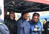 Bald dark-skinned man wearing a blue windbreaker speaks into multiple microphones beneath a tent surrounded by people