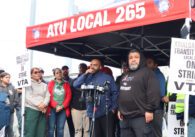 Bald dark-skinned man wearing a blue windbreaker speaks into multiple microphones beneath a red tent labeled "ATU Local 265" with dozens of people beneath it and signs taped to the tent poles reading "Amalgamated Transit Union Local 265 On Strike Against VTA"