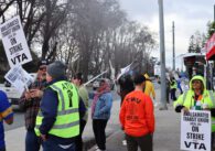 Dozens of people standing on sidewalk, waving signs as cars drive past and talking among themselves. Signs read "Amalgamated Transit Union Local 265 on strike against VTA"