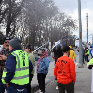 Dozens of people standing on sidewalk, waving signs as cars drive past and talking among themselves. Signs read "Amalgamated Transit Union Local 265 on strike against VTA"