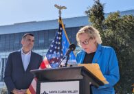 An older woman speaks into a microphone behind a podium while a middle-aged man stands behind her.