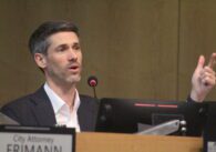 A man sitting at a dais while pointing his finger in the air in front of a microphone.