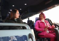 Light skinned woman with dark hair wearing a black puffer jacket sits on an empty bus to the left of a tanner woman with hot pink hair wearing a hot pink jacket