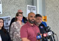 A man speaks into microphones at a podium with people behind him