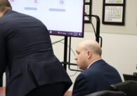 Anthony Becker, white man with light skin and no hair wearing a dark blue suit, sitting at a table beside his lawyers